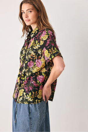 A woman wearing a black short-sleeve button-up shirt with a vibrant multicolor floral print in pink, yellow, and green | FREEMARKET