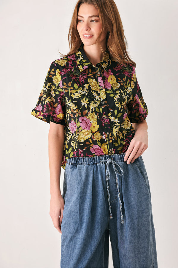 A woman wearing a black short-sleeve button-up shirt with a vibrant multicolor floral print in pink, yellow, and green | FREEMARKET