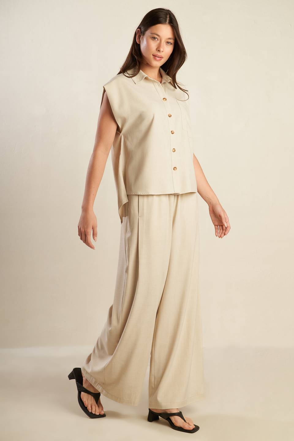 A beige, sleeveless, front button-down, solid woven shirt with collar neck.