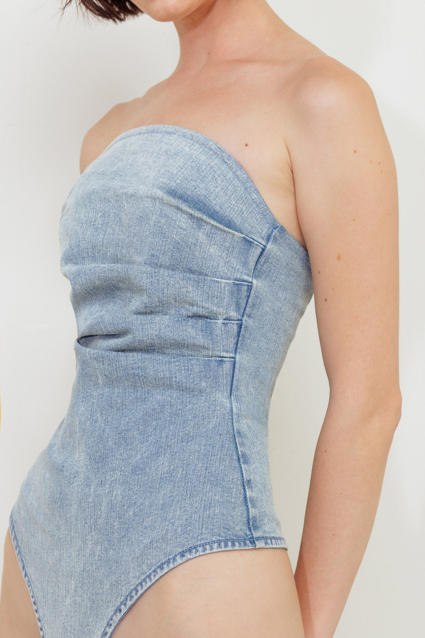 An acid-washed denim bodysuit featuring strapless, side shirring and bottom snap button closure