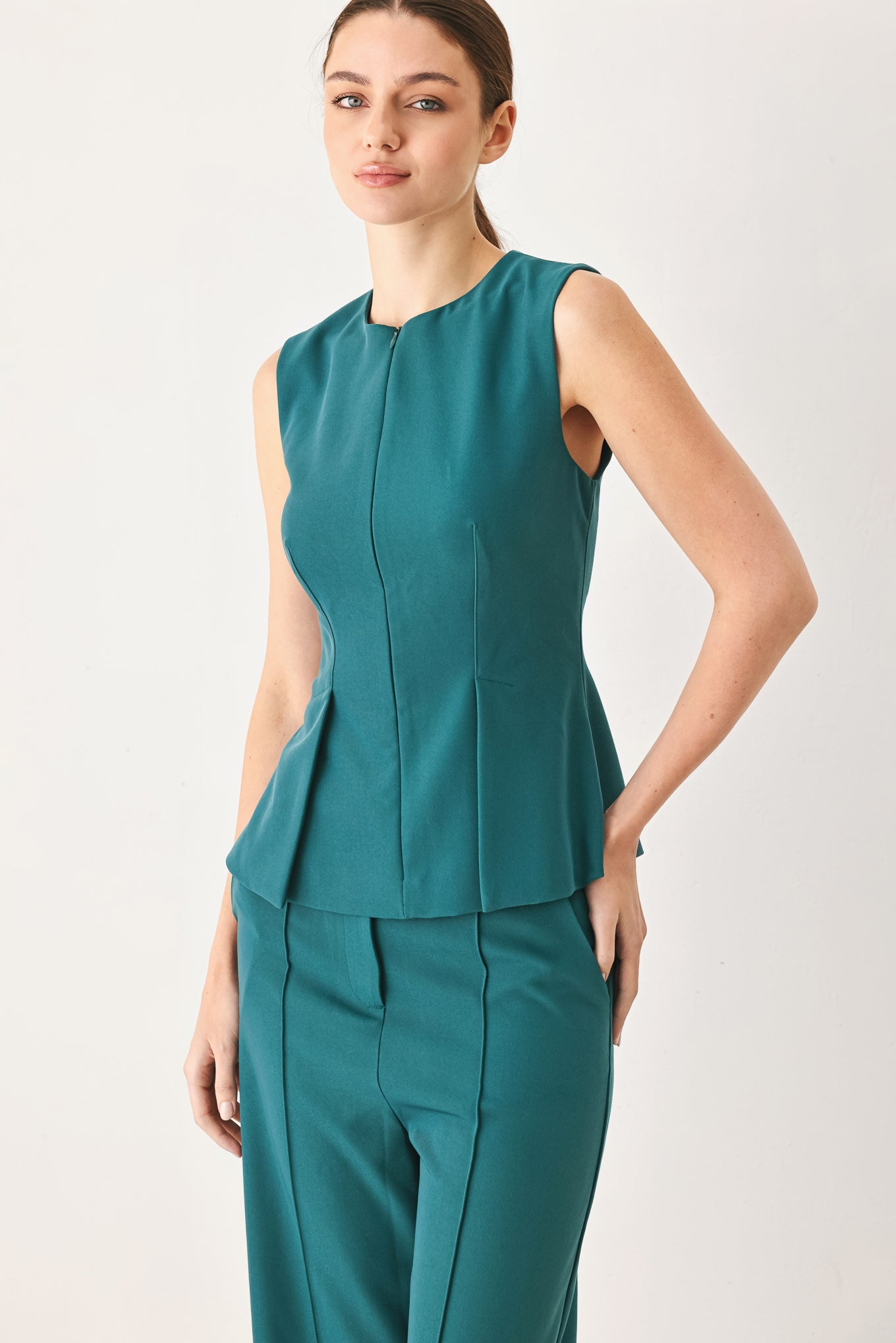 Woman wearing a teal solid woven top featuring round neckline, sleeveless, peplum and front zipper closure | FREEMARKET