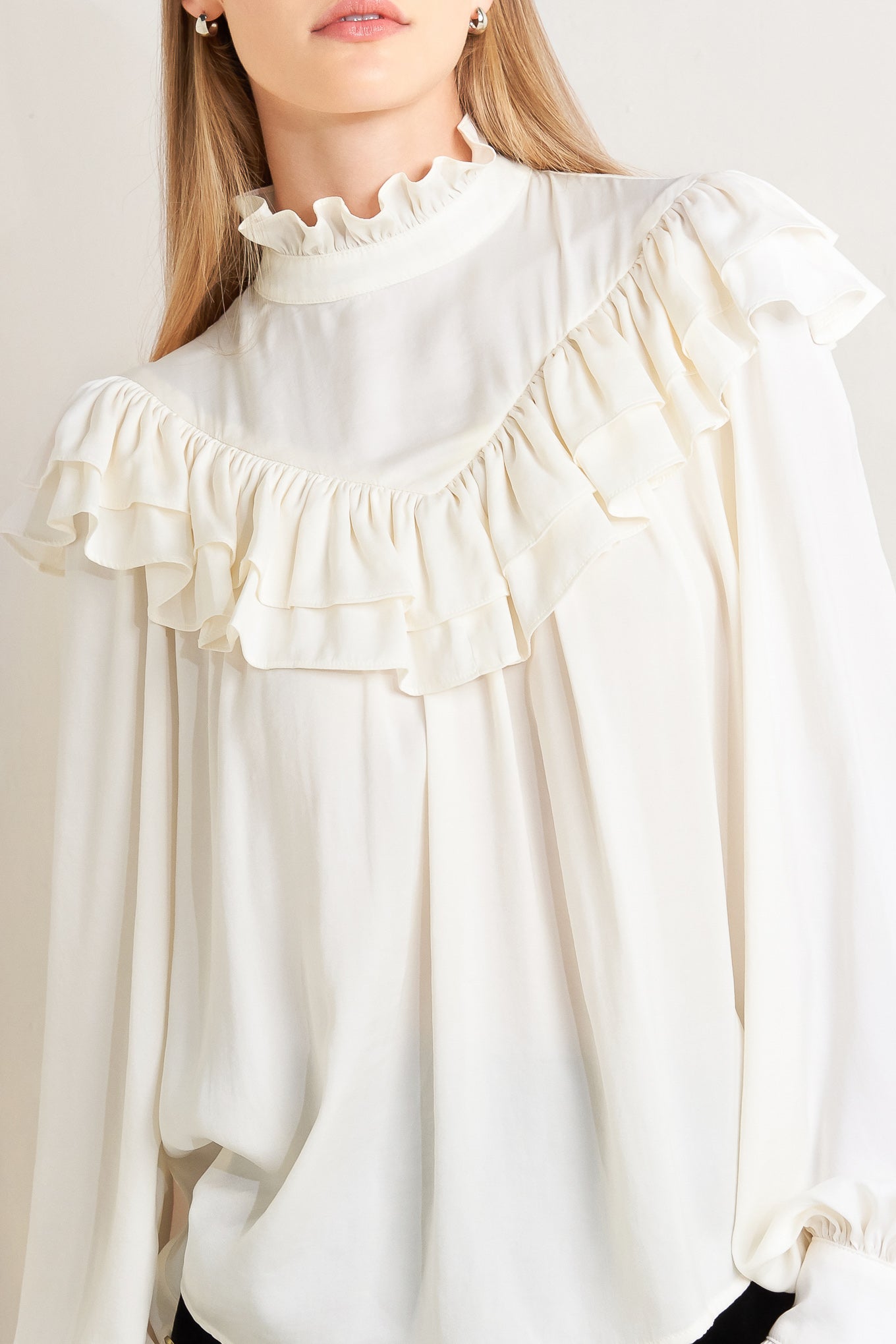 A solid woven top featuring high ruffled neckline, double chevron ruffled front, long sleeve and back neck tie detail