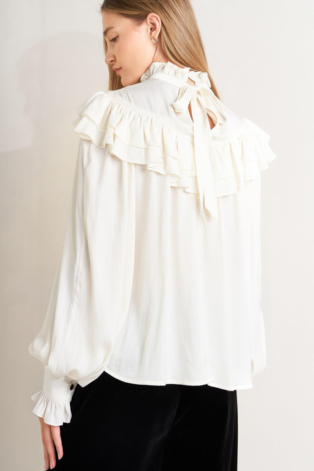 A solid woven top featuring high ruffled neckline, double chevron ruffled front, long sleeve and back neck tie detail