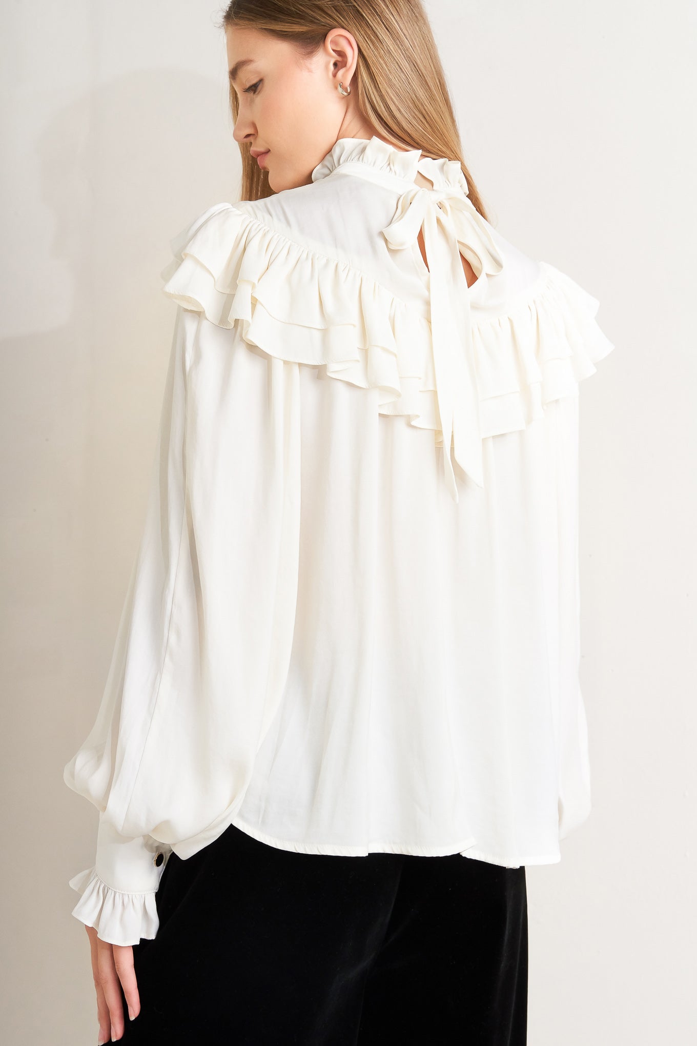 A solid woven top featuring high ruffled neckline, double chevron ruffled front, long sleeve and back neck tie detail