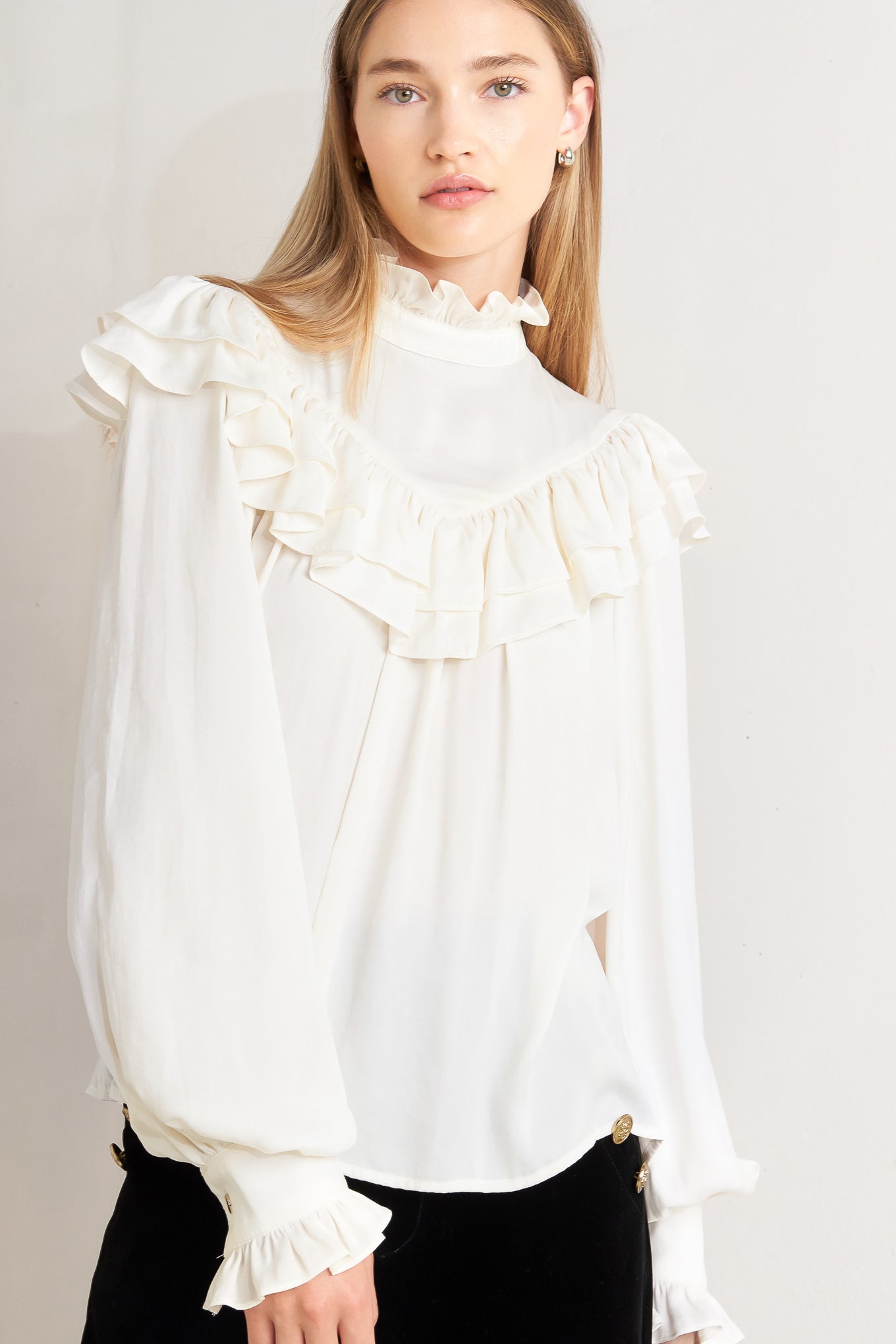 A solid woven top featuring high ruffled neckline, double chevron ruffled front, long sleeve and back neck tie detail