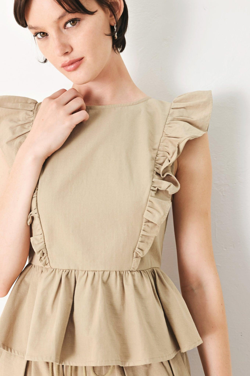 A solid woven top featuring round neckline, ruffle shoulder, peplum, open back with button and tie closure 