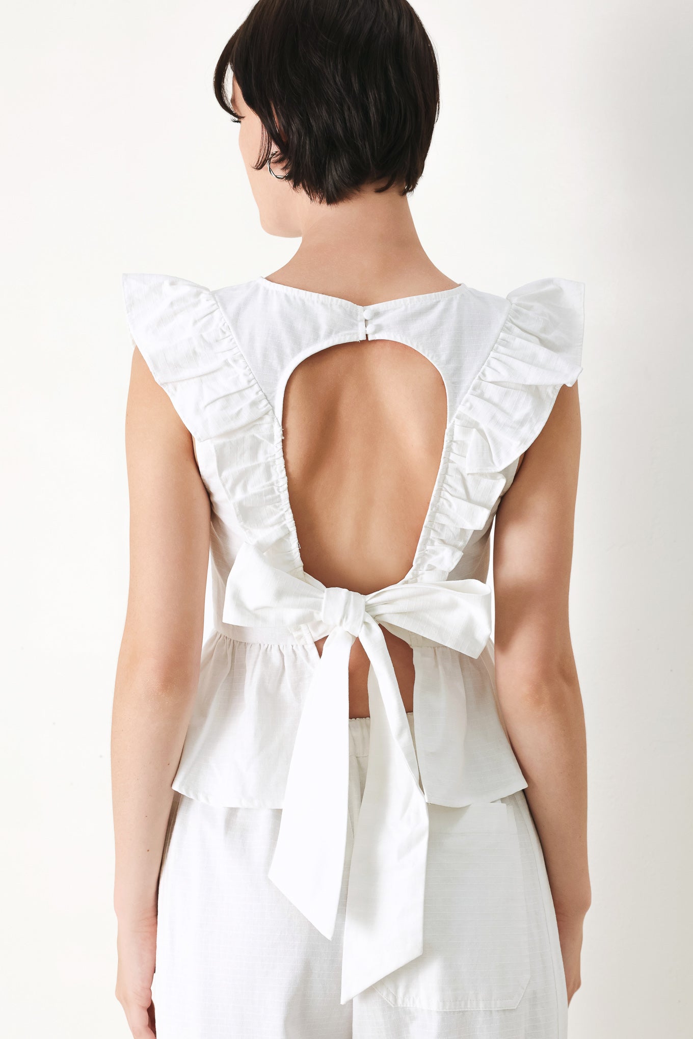 A solid woven top featuring round neckline, ruffle shoulder, peplum, open back with button and tie closure 
