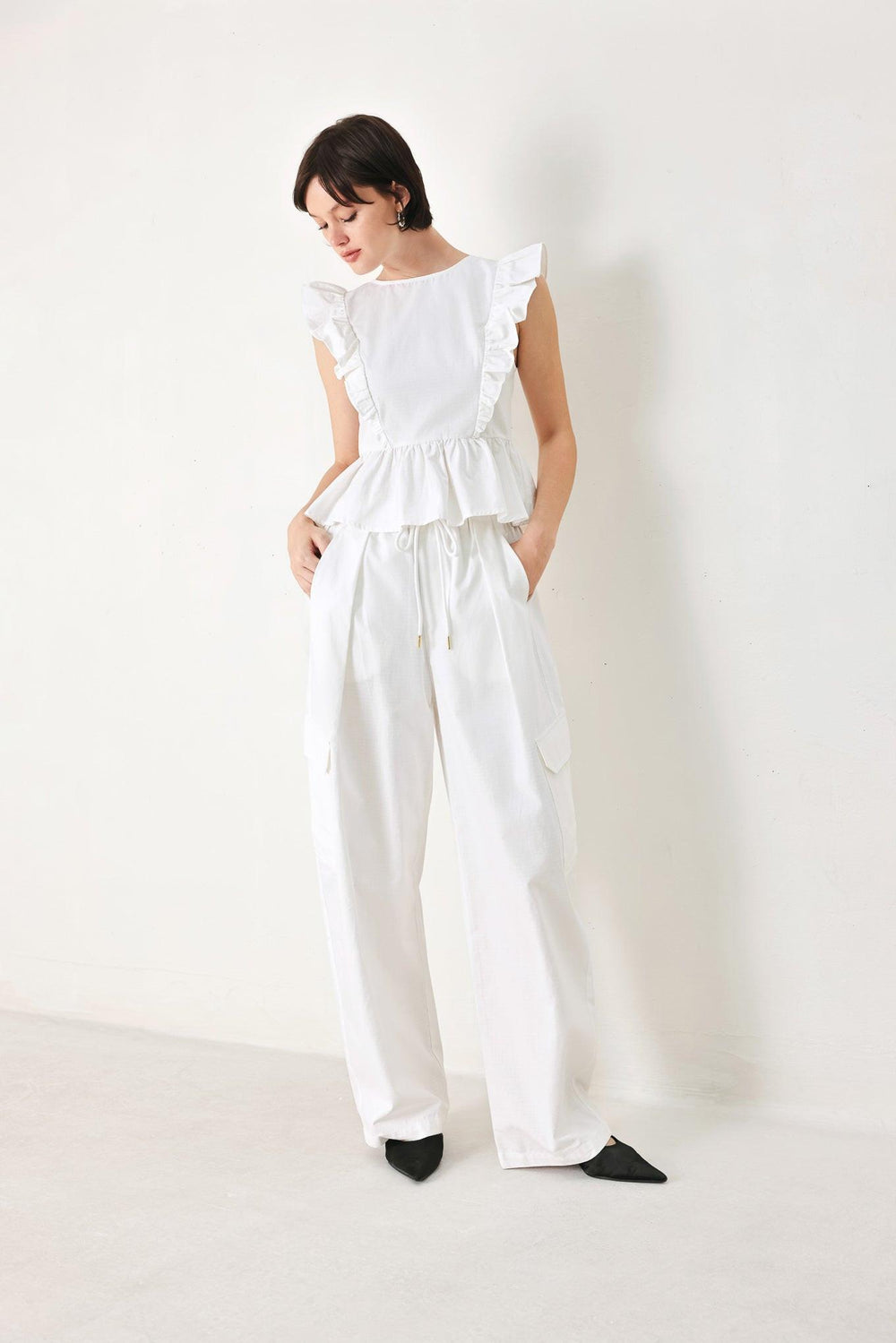 A solid woven top featuring round neckline, ruffle shoulder, peplum, open back with button and tie closure 