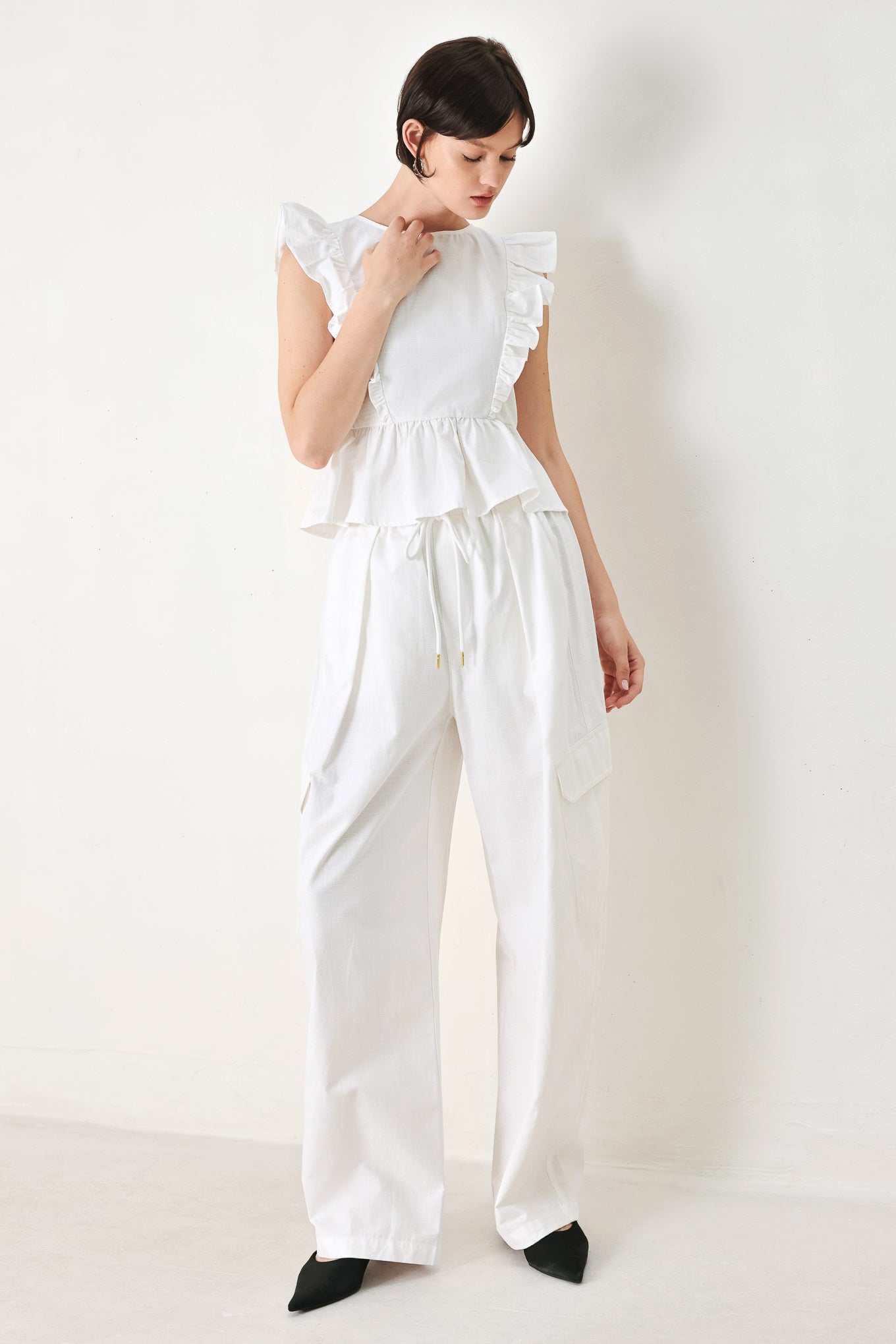 A solid woven top featuring round neckline, ruffle shoulder, peplum, open back with button and tie closure 