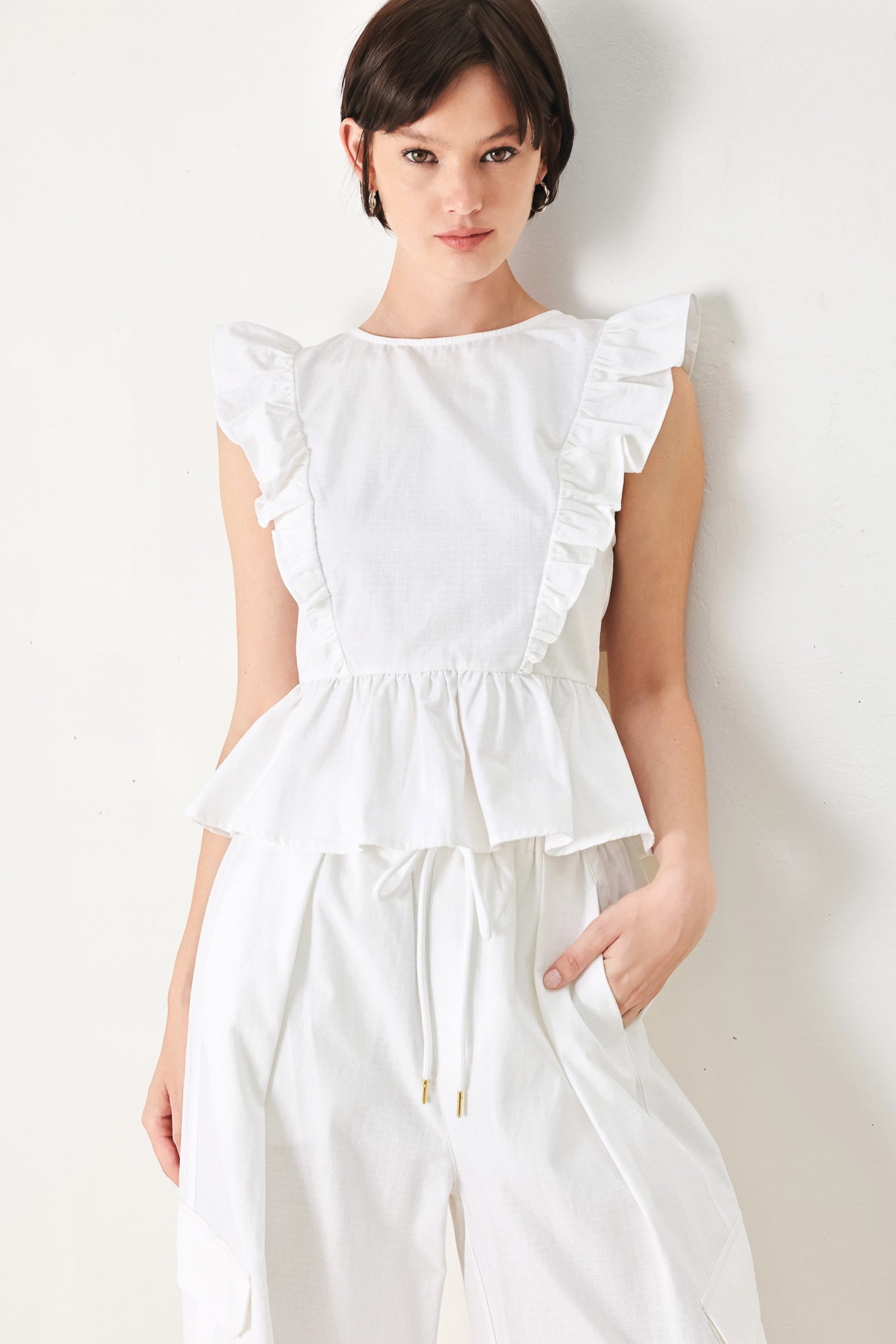A solid woven top featuring round neckline, ruffle shoulder, peplum, open back with button and tie closure 