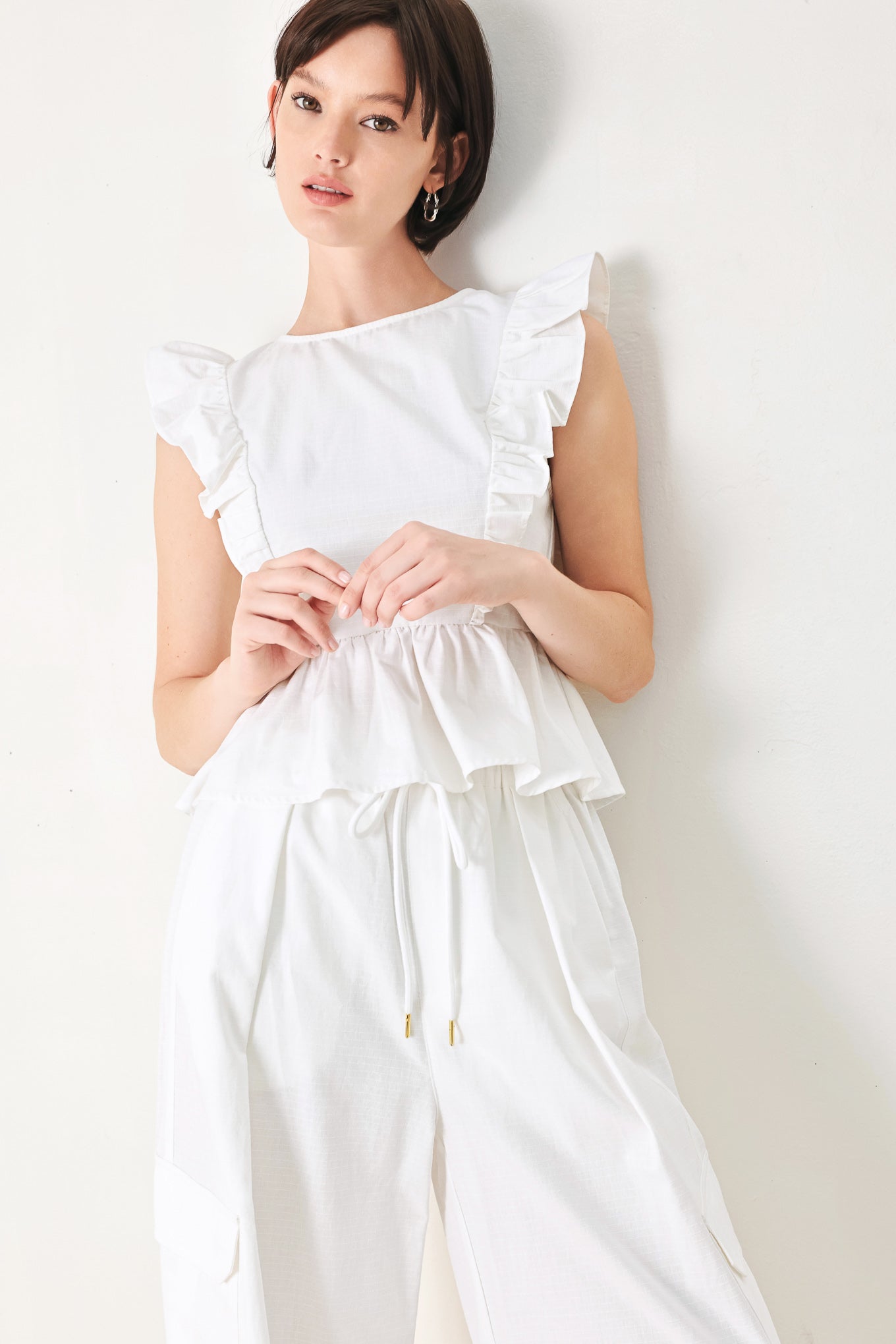 A solid woven top featuring round neckline, ruffle shoulder, peplum, open back with button and tie closure 