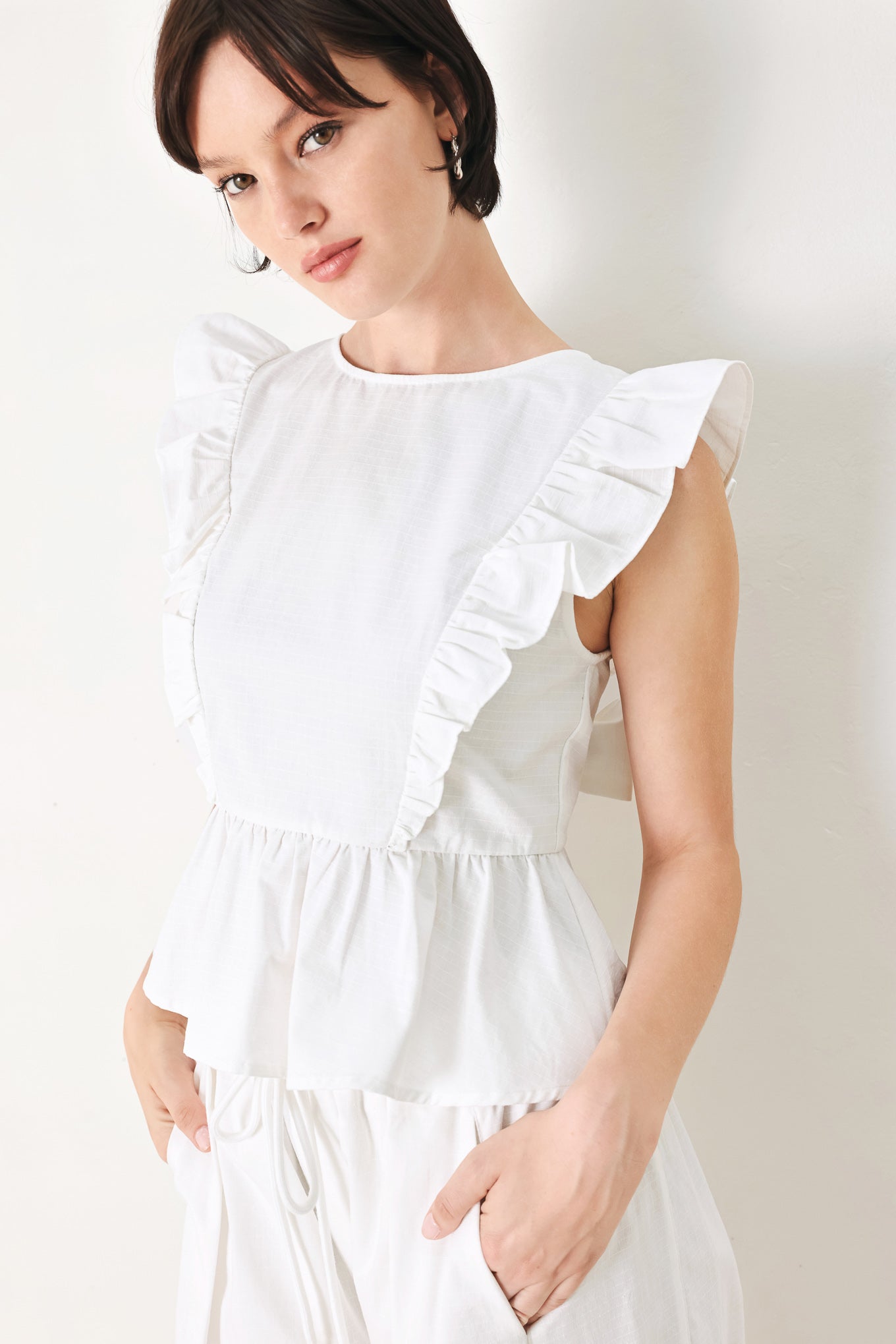 A solid woven top featuring round neckline, ruffle shoulder, peplum, open back with button and tie closure 