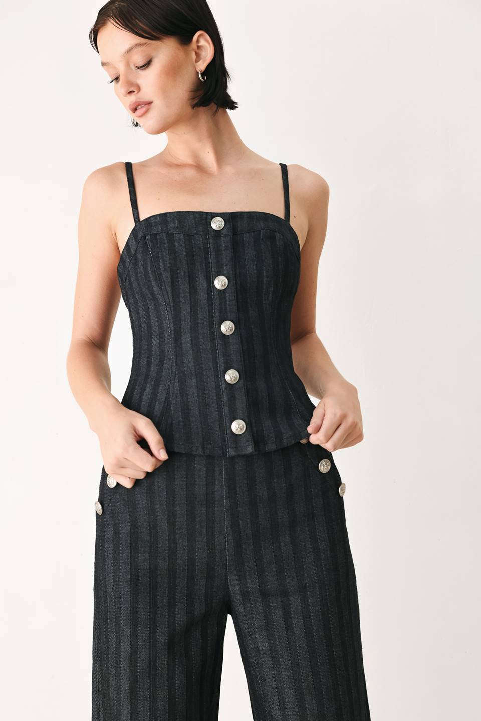 A model wearing a dark gray pinstriped jumpsuit with a structured corset-style bodice, thin shoulder straps, and prominent silver-tone decorative buttons down the front and on the waist | FREEMARKET