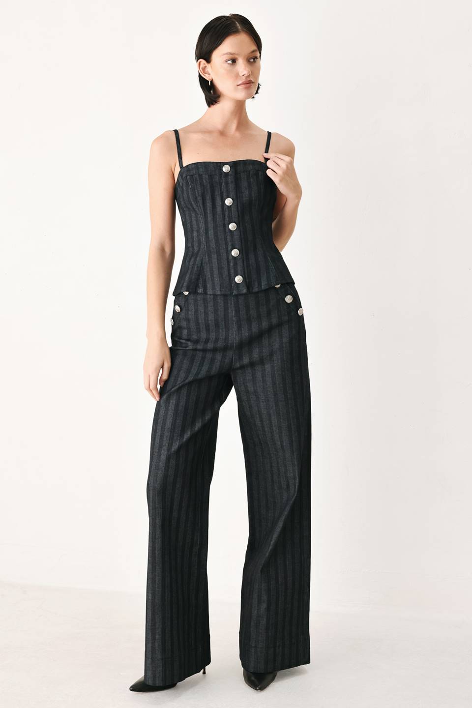 A model wearing a dark gray pinstriped jumpsuit with a structured corset-style bodice, thin shoulder straps, and prominent silver-tone decorative buttons down the front and on the waist | FREEMARKET