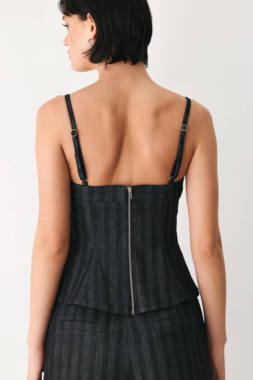 A model wearing a dark gray pinstriped jumpsuit with a structured corset-style bodice, thin shoulder straps, and prominent silver-tone decorative buttons down the front and on the waist | FREEMARKET
