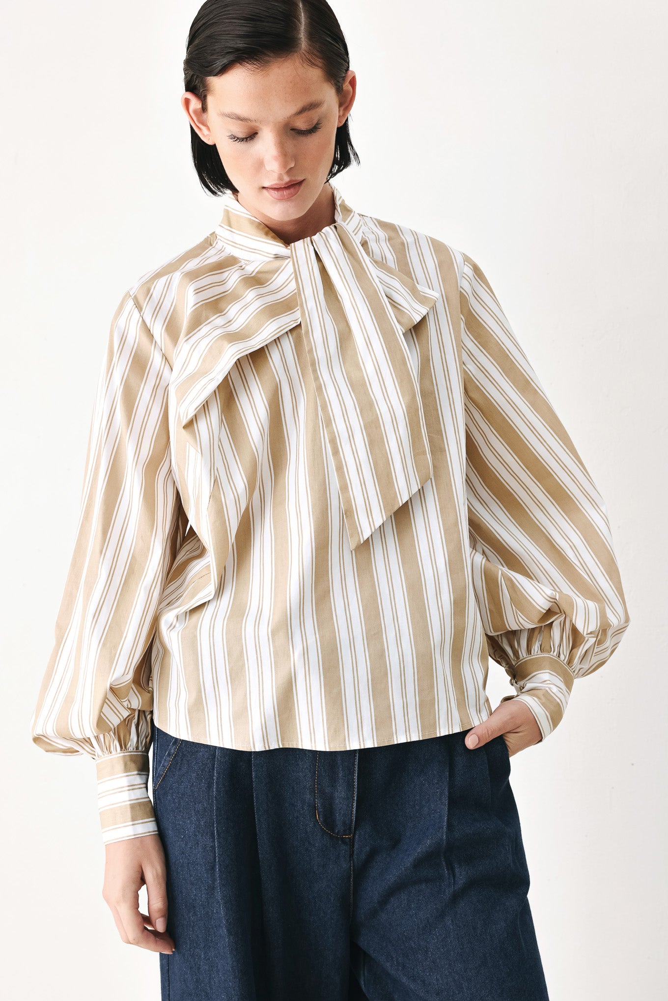 A model wearing a khaki beige and white vertically striped blouse with a large ruffled bow at the collar and voluminous long sleeves | FREEMARKET