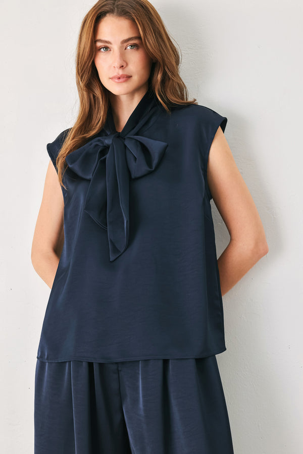 Woman wearing a navy blue cap sleeve satin top with a bow along the neck | FREEMARKET