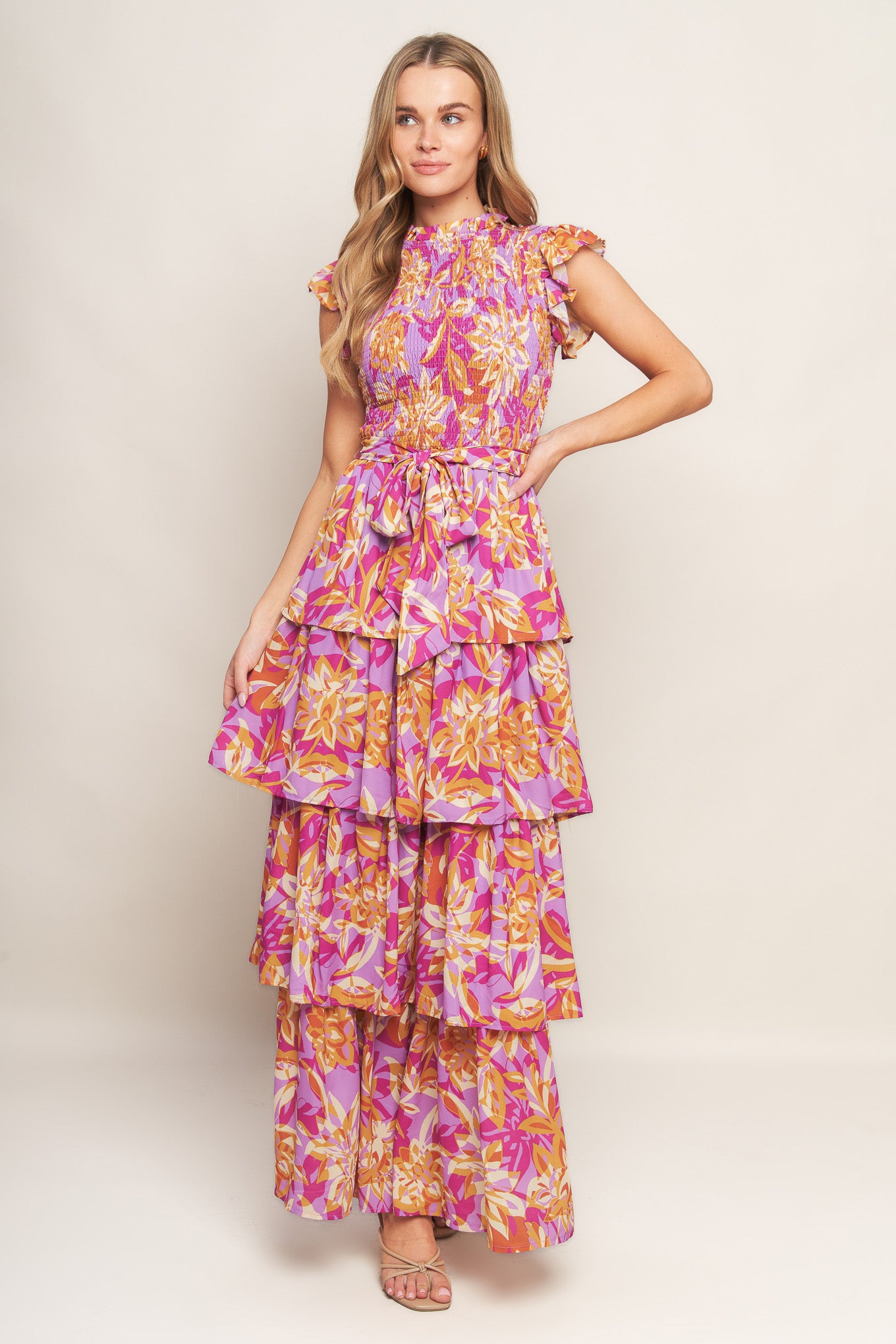 A printed woven midi dress featuring ruffled neckline, sleeveless with ruffled edge, smocked bodice, self belt, layered skirt and back neck button closure