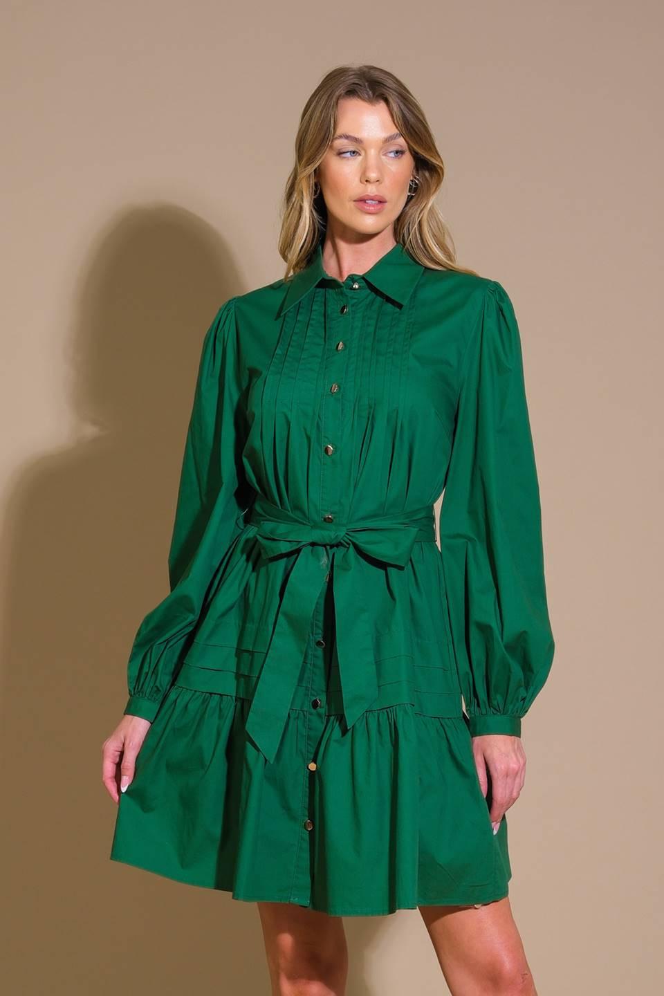 A solid woven mini dress featuring a shirt collar, button down, front pleats, long sleeve, self-sash tie, and ruffled hemline