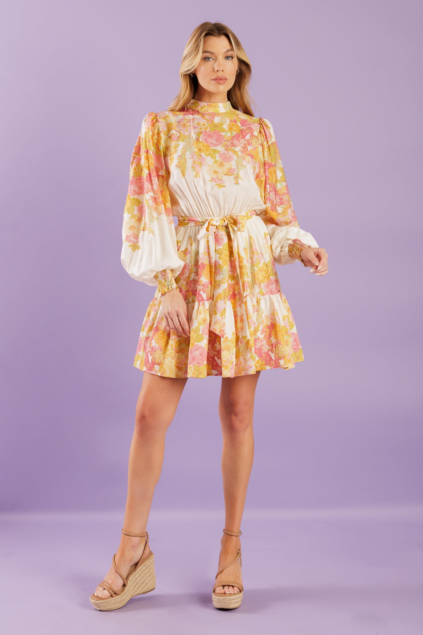 A printed woven mini dress featuring high neckline, long sleeve with smocked cuff, elasticized waist, ruffled hemline and back neck button closure.