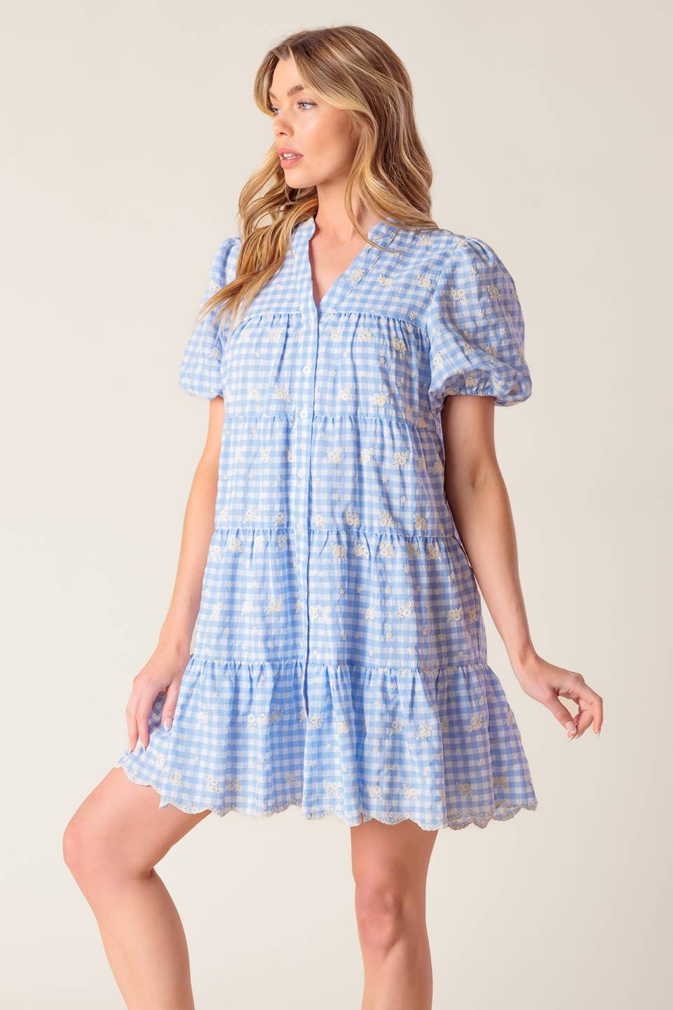 A woven powder blue gingham babydoll dress featuring V neckline, short puff sleeve and tiered body with ruffled hemline.