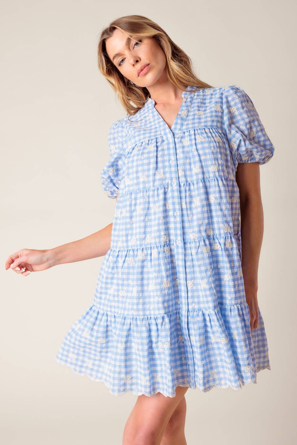 A woven powder blue gingham babydoll dress featuring V neckline, short puff sleeve and tiered body with ruffled hemline.