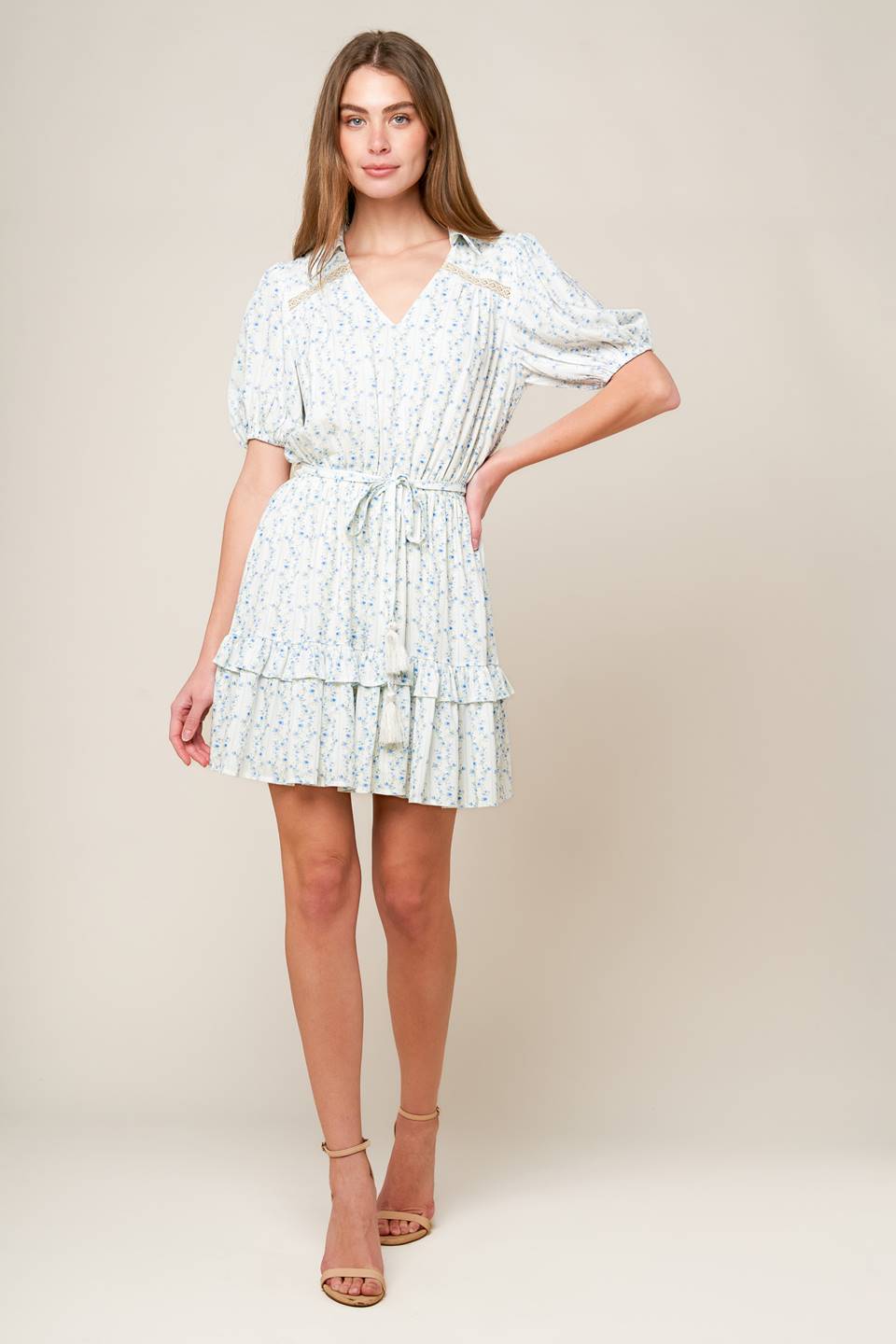 A white woven mini dress featuring blue printed florals, V neckline with collar, short puff sleeve, front and back lace inset, elasticized waist with self sash tie and ruffled hemline.