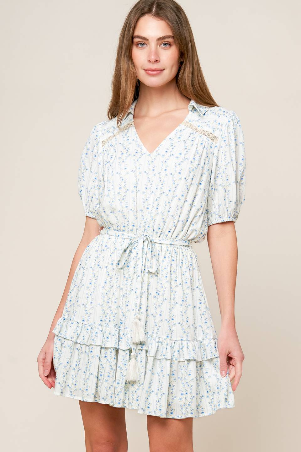 A white woven mini dress featuring blue printed florals, V neckline with collar, short puff sleeve, front and back lace inset, elasticized waist with self sash tie and ruffled hemline.