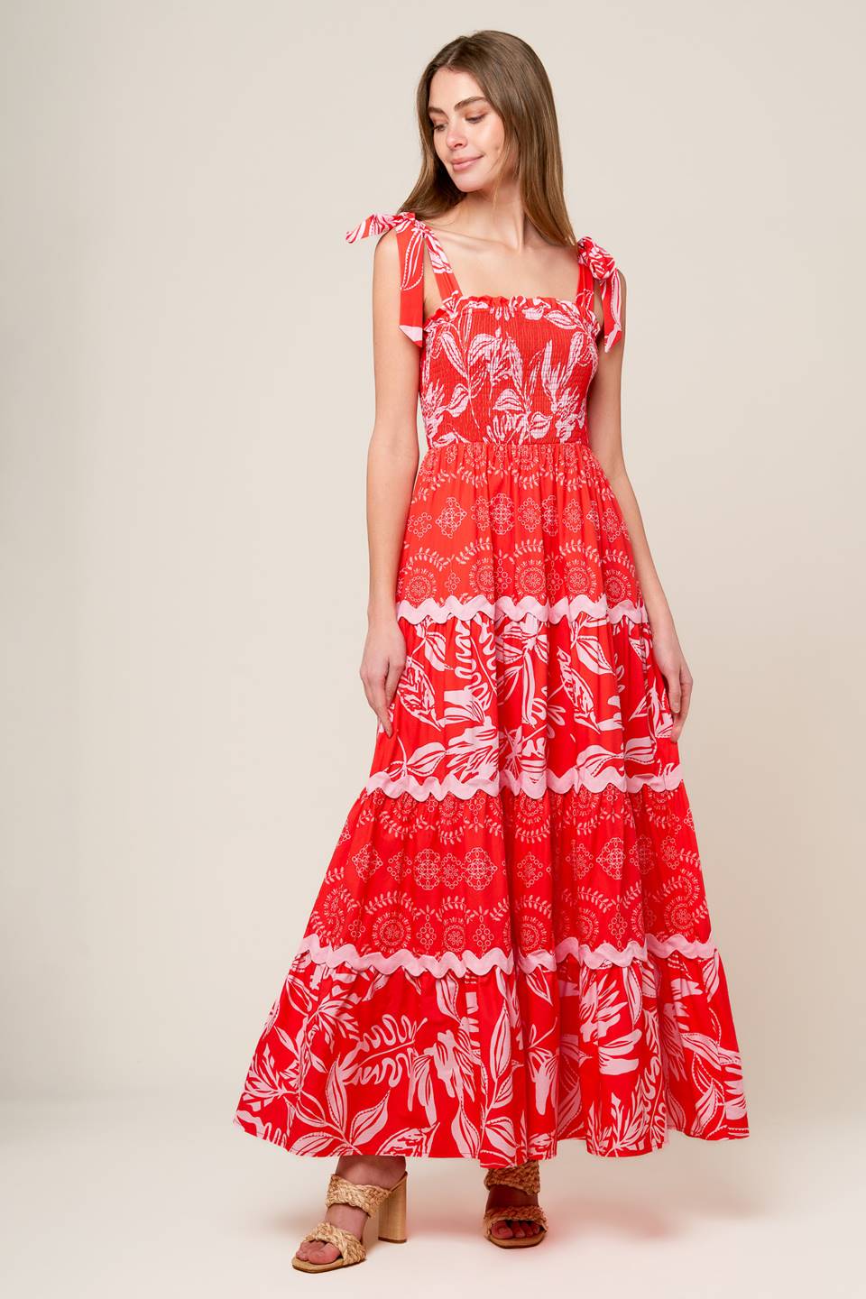 A red and pink printed woven maxi dress featuring straight neckline with ruffled edge, shoulder ties, smocked bodice and tiered skirt with ric-rac ribbon inset.