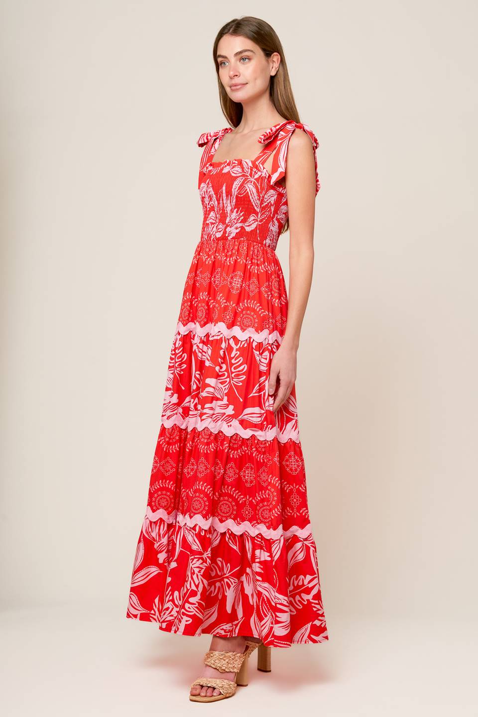 A red and pink printed woven maxi dress featuring straight neckline with ruffled edge, shoulder ties, smocked bodice and tiered skirt with ric-rac ribbon inset.