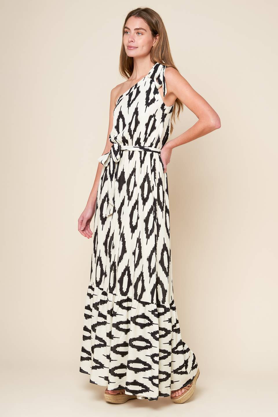 A printed woven maxi dress featuring one shoulder neckline with tie, elasticized waist with self sash tie and ruffled hemline.