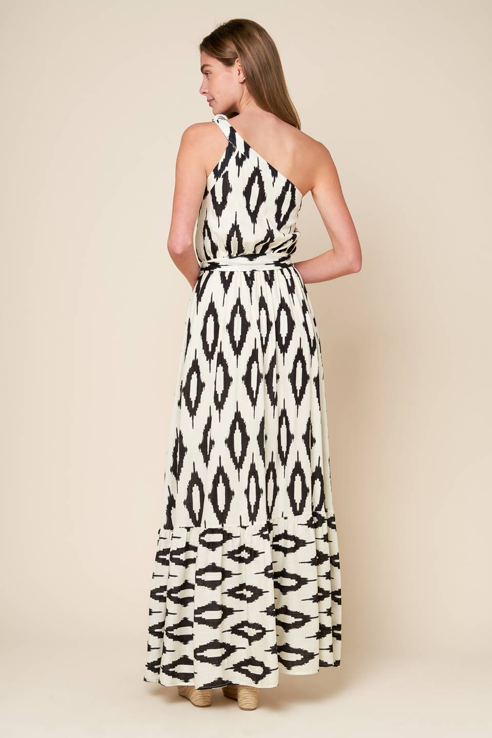 A printed woven maxi dress featuring one shoulder neckline with tie, elasticized waist with self sash tie and ruffled hemline.