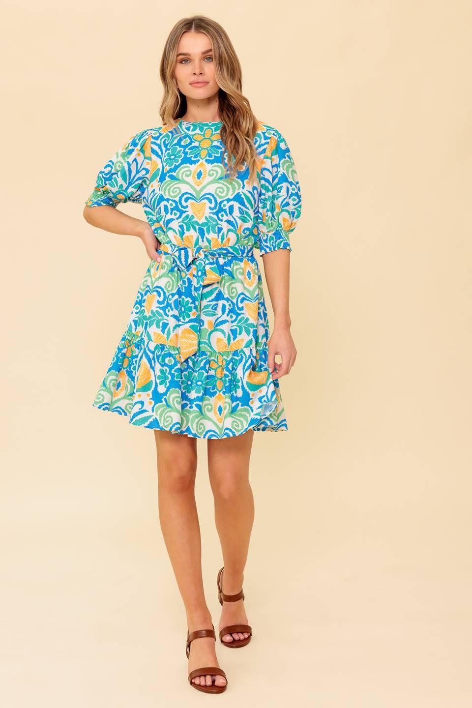 A printed woven mini dress featuring round neckline, short puff sleeve with smocked cuff, elasticized waist with self sash tie, ruffled hemline and back neck button closure