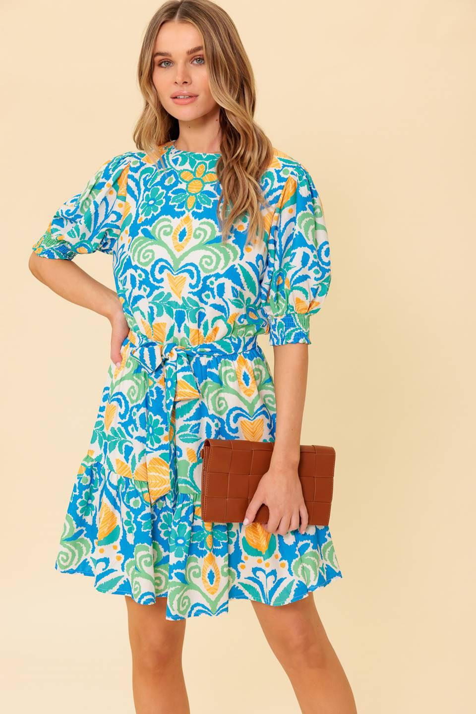 A printed woven mini dress featuring round neckline, short puff sleeve with smocked cuff, elasticized waist with self sash tie, ruffled hemline and back neck button closure