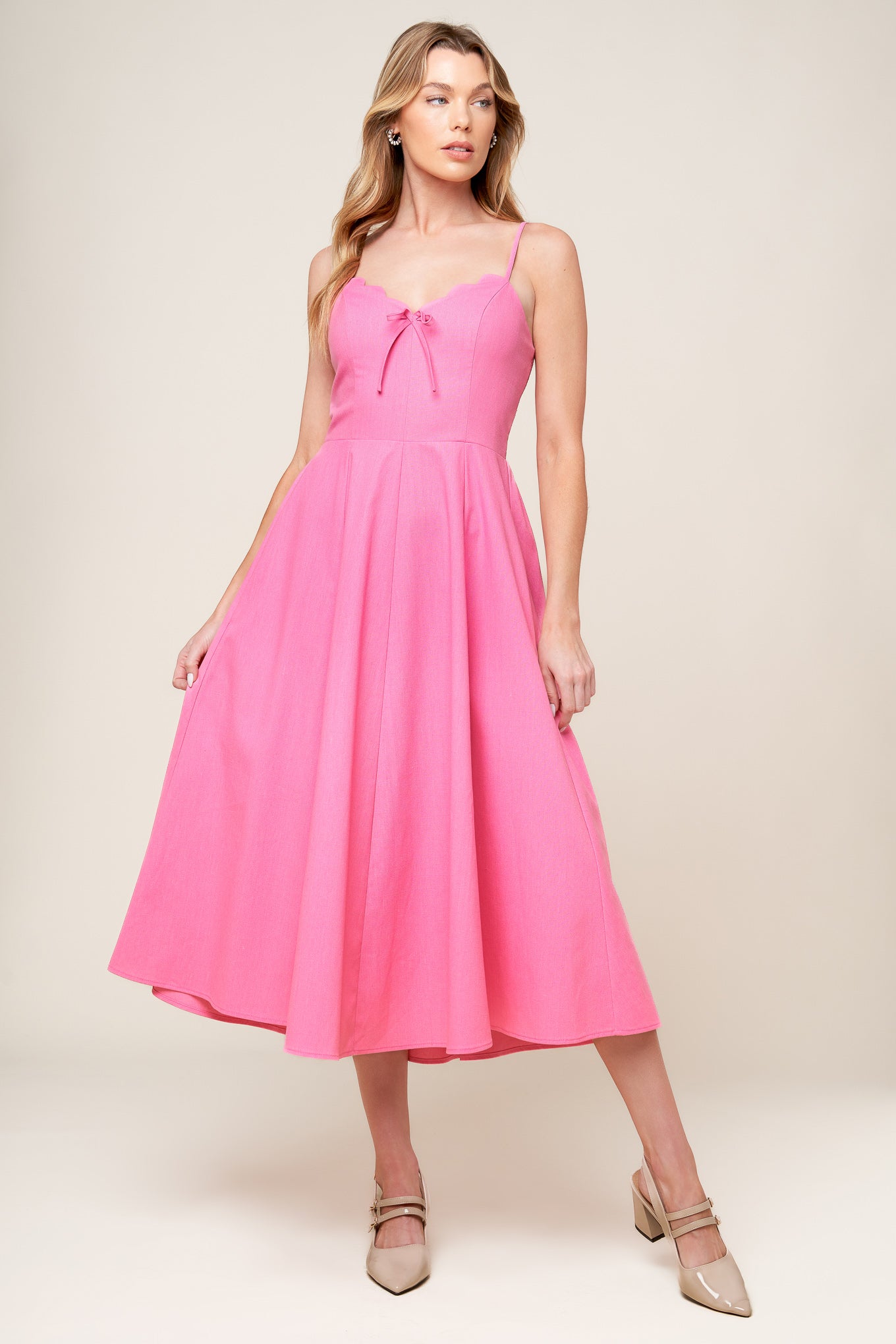 A solid pink woven midi dress featuring V neckline with scalloped edge, strap, front bow tie detail, circle skirt and smocked back bodice.