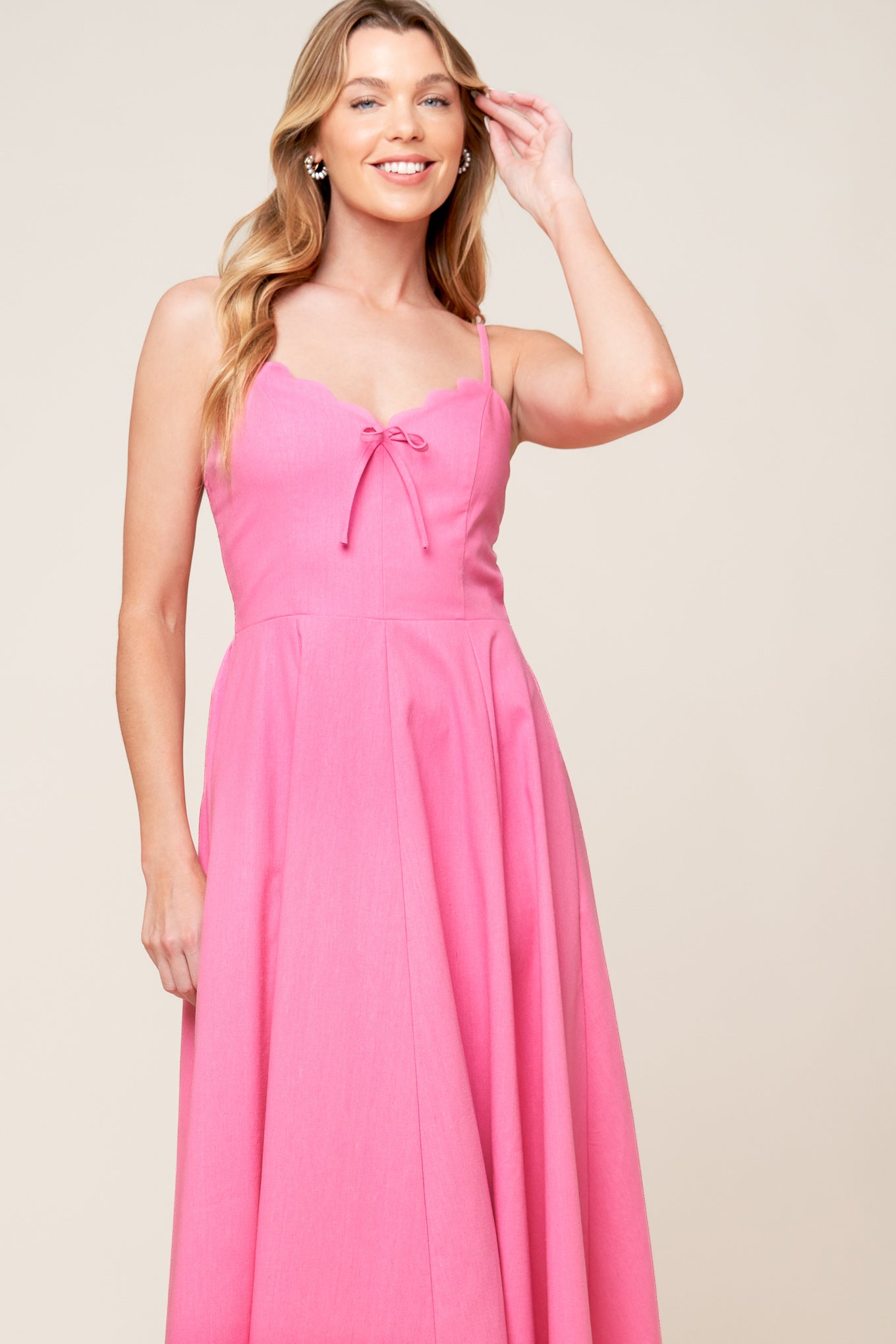 A solid pink woven midi dress featuring V neckline with scalloped edge, strap, front bow tie detail, circle skirt and smocked back bodice.