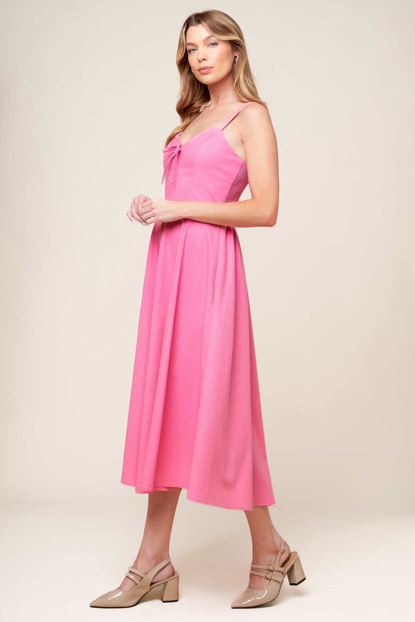 A solid pink woven midi dress featuring V neckline with scalloped edge, strap, front bow tie detail, circle skirt and smocked back bodice.