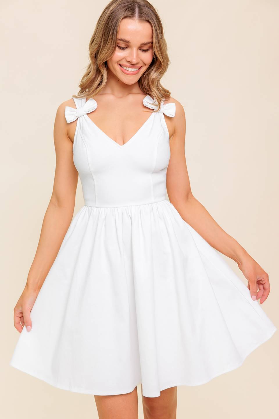 A solid white woven mini dress featuring V-neckline, straps with bow, full skirt and back zipper closure