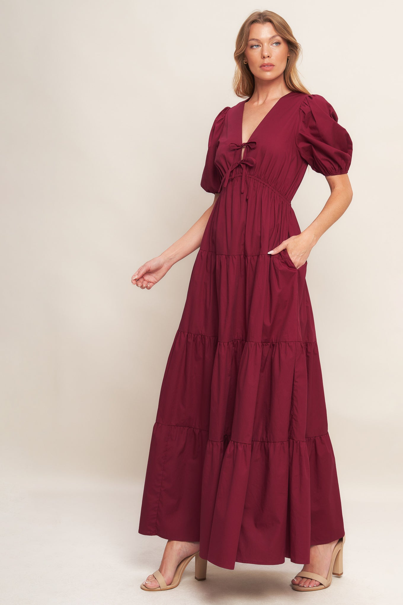 A solid woven maxi dress featuring V neckline with front bow closure, short puff sleeve, elasticized empire and tiered skirt.