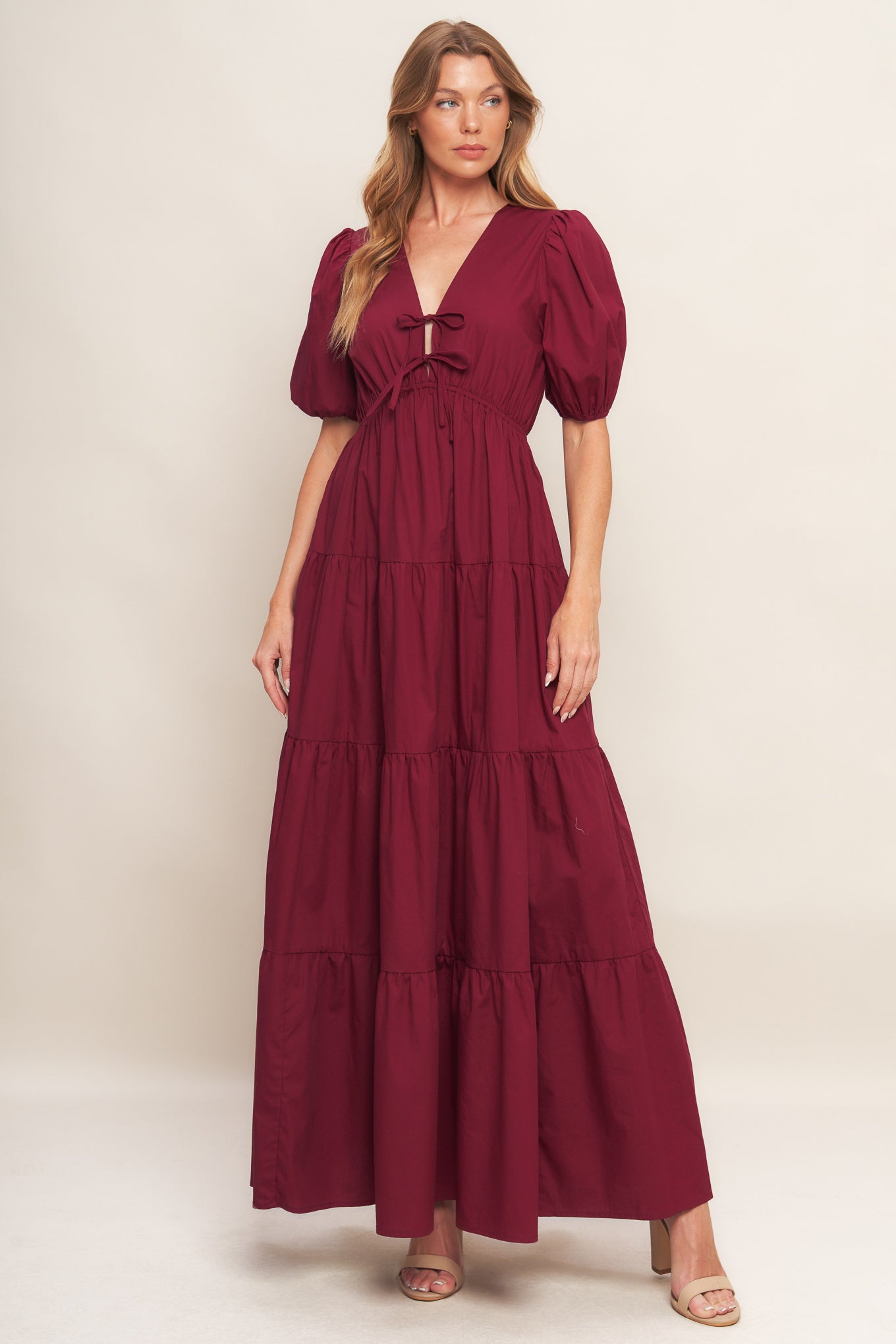 A solid woven maxi dress featuring V neckline with front bow closure, short puff sleeve, elasticized empire and tiered skirt.
