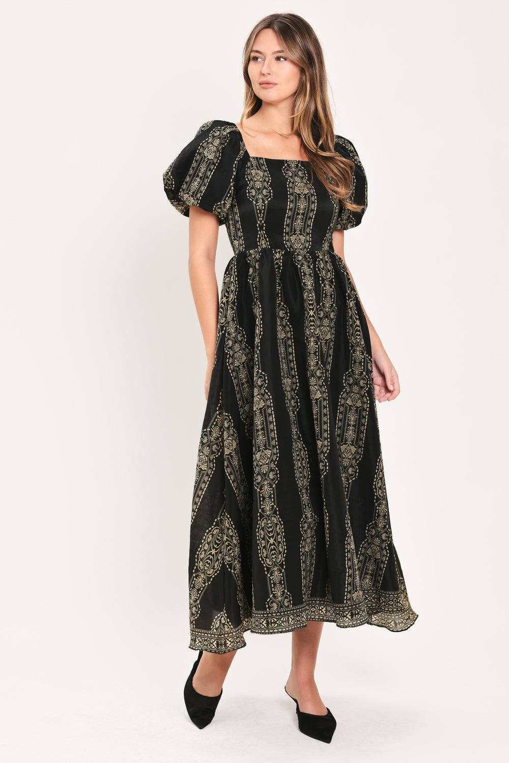 A woman modeling a black midi dress with a beige geometric pattern, puff sleeves, and a square neckline, paired with black pointed-toe mule heels against a plain light background.