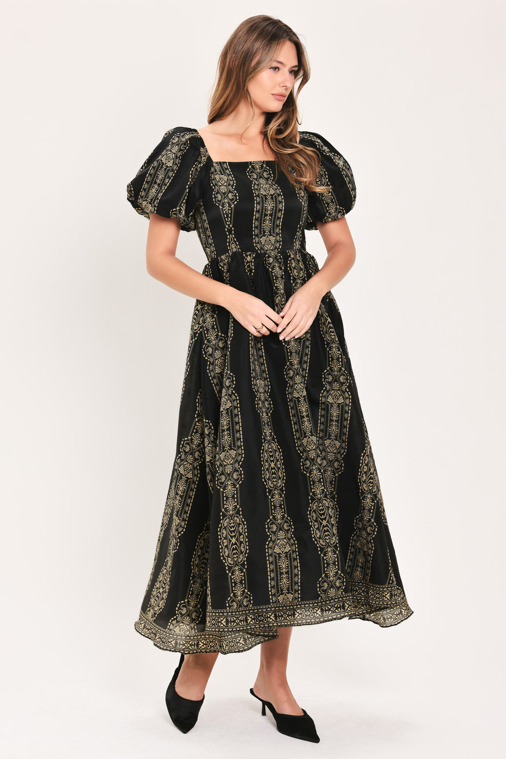 A woman modeling a black midi dress with a beige geometric pattern, puff sleeves, and a square neckline, paired with black pointed-toe mule heels against a plain light background.