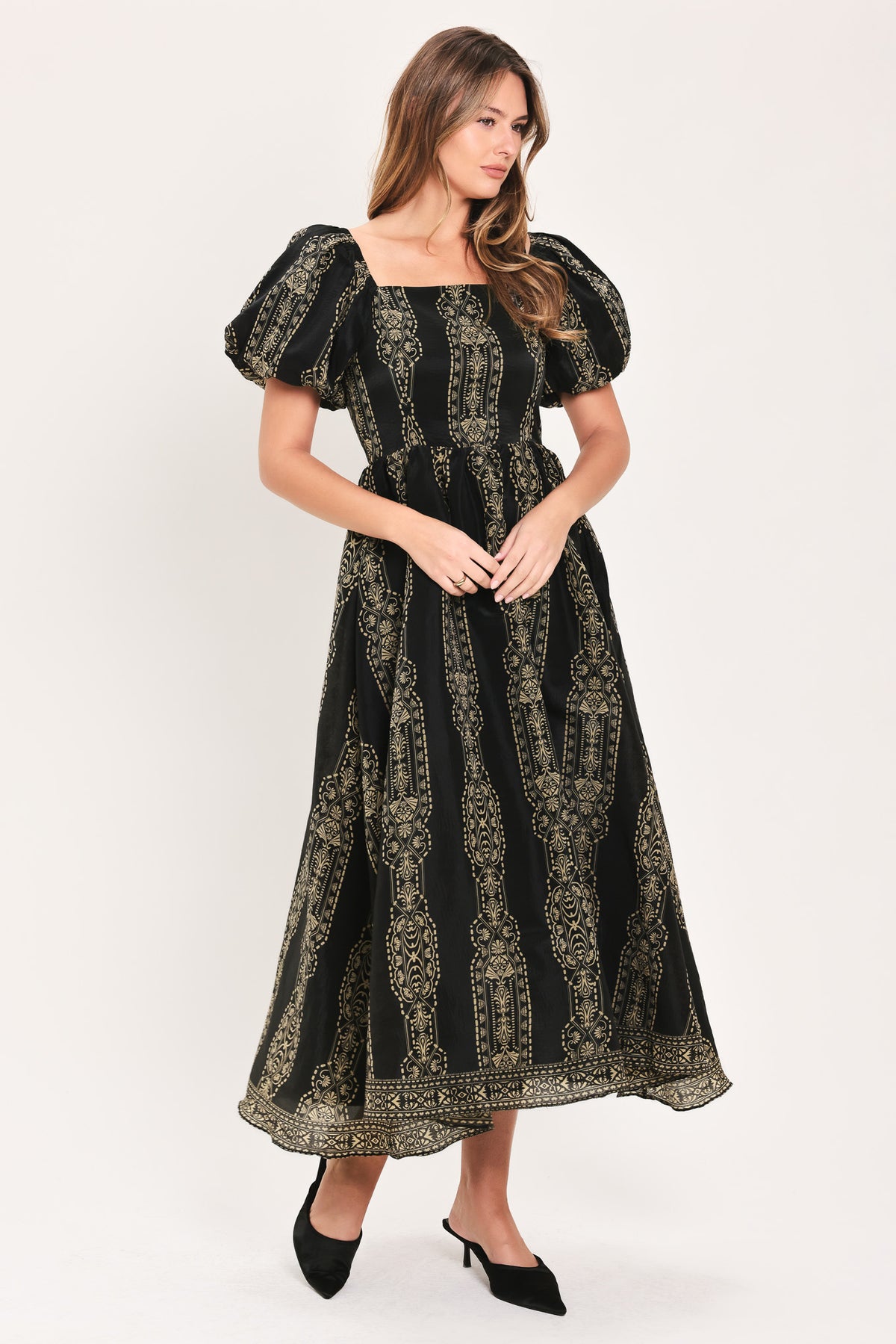 A woman modeling a black midi dress with puff sleeves and an intricate gold vertical pattern, paired with black pointed-toe heels, against a plain light gray studio background.