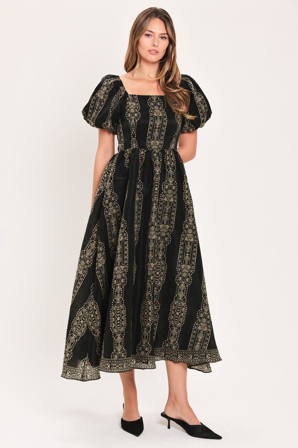A woman modeling a black midi dress with a beige geometric pattern, puff sleeves, and a square neckline, paired with black pointed-toe mule heels against a plain light background.