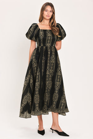 A woman modeling a black midi dress with a beige geometric pattern, puff sleeves, and a square neckline, paired with black pointed-toe mule heels against a plain light background.