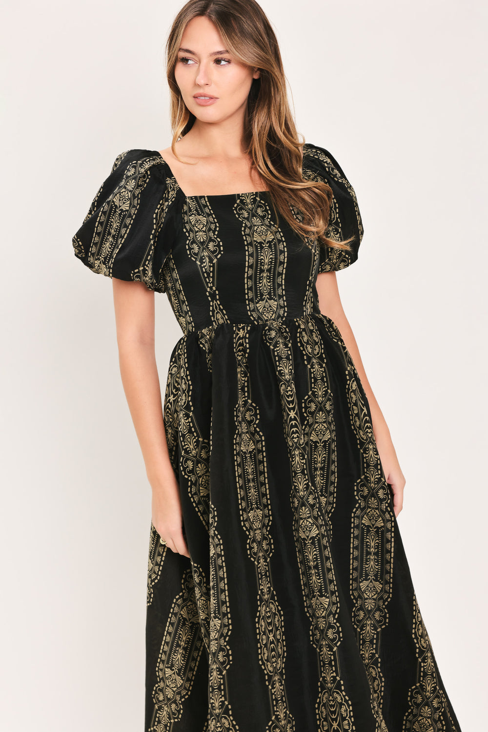 A woman modeling a black midi dress with a beige geometric pattern, puff sleeves, and a square neckline, paired with black pointed-toe mule heels against a plain light background.
