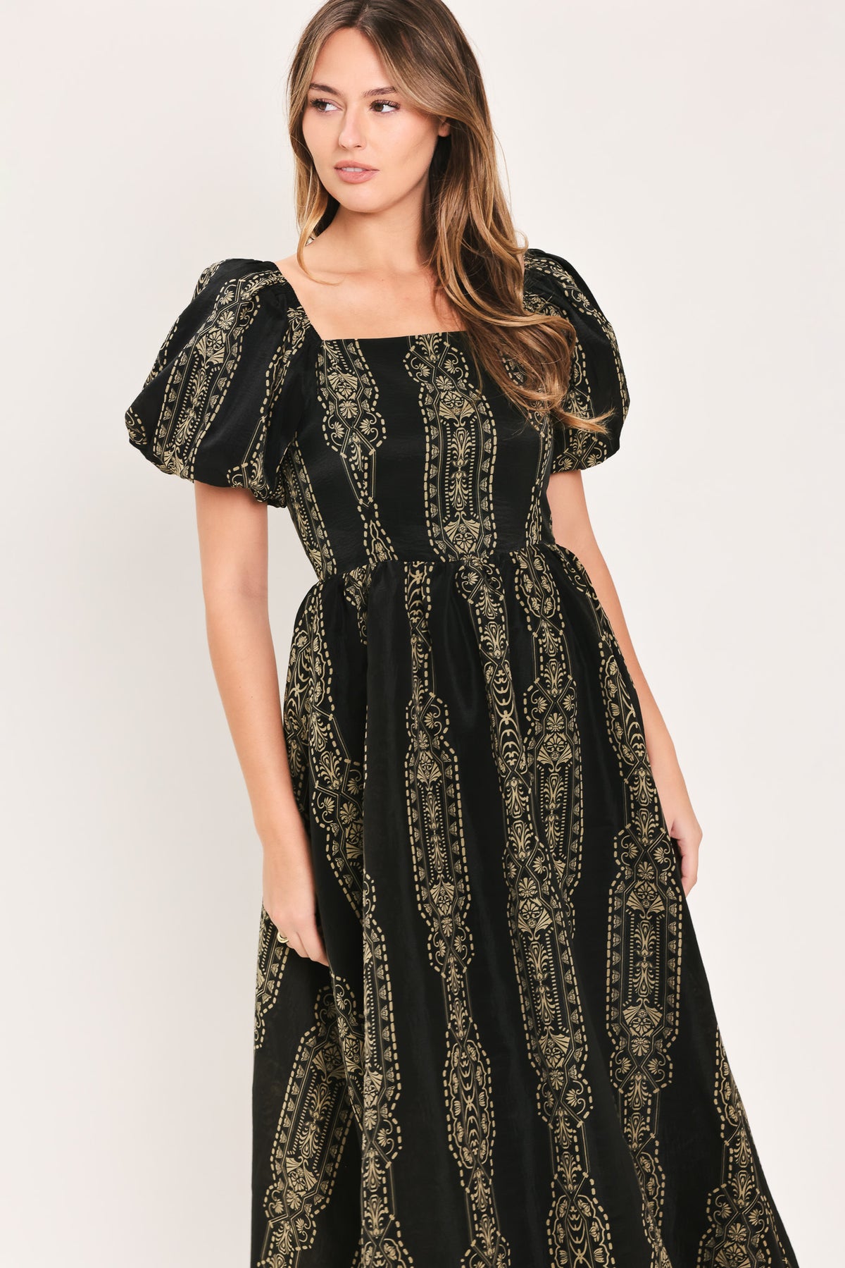 A woman wearing a black midi dress with puff sleeves and a gold geometric pattern, photographed against a plain light background.