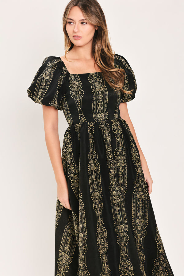 A woman wearing a black midi dress with puff sleeves and a gold geometric pattern, photographed against a plain light background.