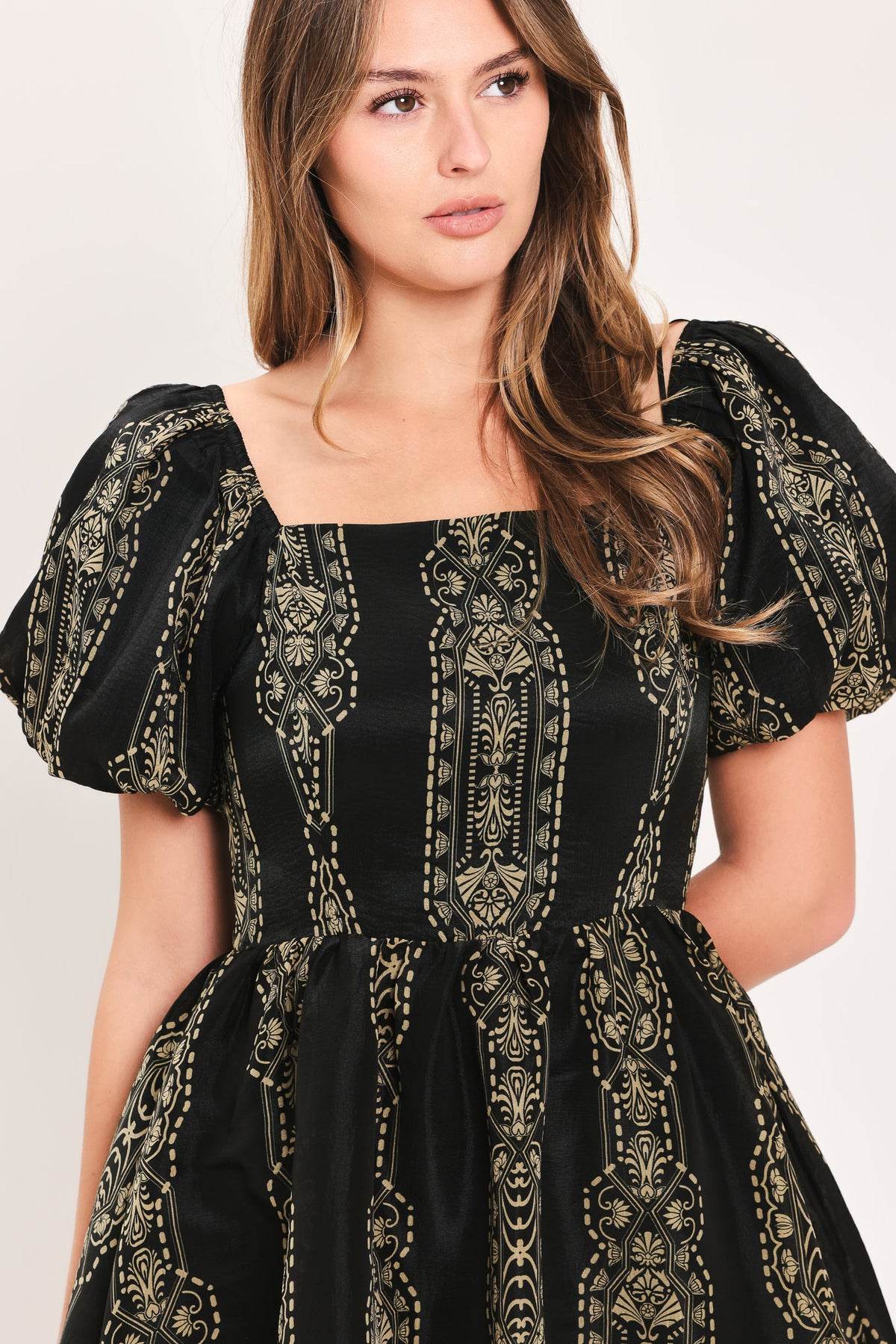 A woman wearing a black dress with ornate gold and beige bohemian-style embroidery, featuring puff sleeves and a square neckline, set against a plain light background.