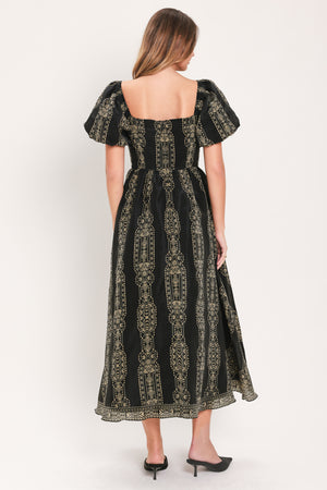 A woman viewed from the back wearing a black midi dress with puff sleeves and an intricate vertical gold pattern, paired with black pointed-toe heels against a plain white studio background.